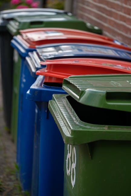 A residential street scene showing a woman standing next to a row of large black wheelie bins positioned along the pavement in front of red-brick terraced houses with white window frames. The bins are