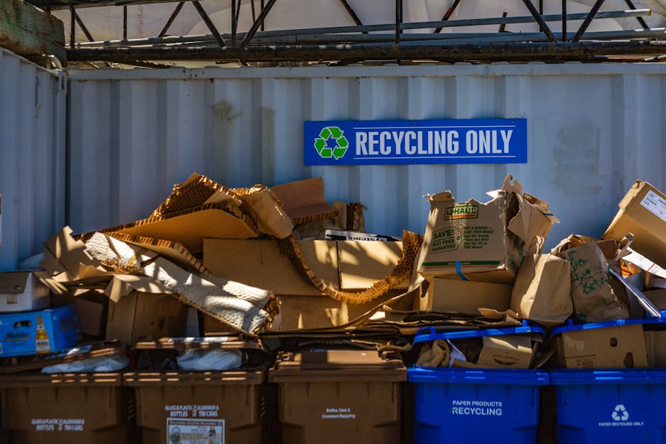 A large collection of flattened cardboard boxes, some torn or crumpled, are stacked together in front of a grey metal wall with a blue and green 'Recycling Only' sign clearly visible above. The boxes 