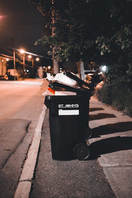 A sanitation worker wearing a high-visibility yellow vest and dark trousers is operating a red waste collection vehicle parked on the side of a street. The back of the vehicle is open, revealing mecha